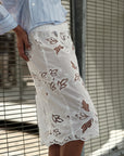A person stands by a wire fence wearing Cissa's The Summer Pencil Skirt, featuring intricate cut-out floral embroidery and a scalloped hem, seen from waist to knees.