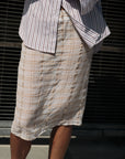 A person stands outdoors in a Cissa Summer Pencil Skirt, a beige pull-on midi with an elastic waistband, paired with an untucked white shirt with vertical maroon stripes; metal bars are visible in the background.