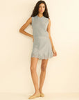 A woman with long dark hair wears DOEN's Giulietta Skirt— a sleeveless light gray knit mini skirt with a floral lace hem—paired with a matching top and beige flats, standing against a beige background.