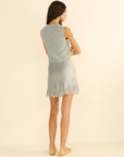 A woman with long dark hair, barefoot against a plain light background, models the DOEN Giulietta Skirt—a matching silk satin mini skirt with a floral lace hem—paired with a sleeveless light blue knit top.