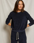 A woman with straight, center-parted brown hair stands against a light beige backdrop, wearing the Perfectwhitetee Saylor Loop Terry Sweatshirt in dark navy with matching drawstring pants. Her hands rest on her hips.