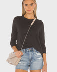 A woman stands facing forward against a plain white background, wearing The LS Slim Tee by The Great Inc., light blue denim shorts, and a beige crossbody purse.