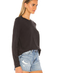 A woman wears The LS Slim Tee by The Great Inc., a black long-sleeve cotton jersey top, with light blue distressed denim shorts, standing sideways against a white background.