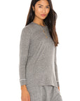 A woman with long, dark hair and gold jewelry smiles slightly against a white background, wearing The Great Inc.'s The LS Slim Tee—a grey cotton jersey long-sleeve top—paired with matching pants.