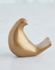 The Les Animaux Moineau Gold sculpture by Global Views features a smooth, stylized bird with a rounded body and curved tail in Matte Antique Gold, displayed on a light gray surface.
