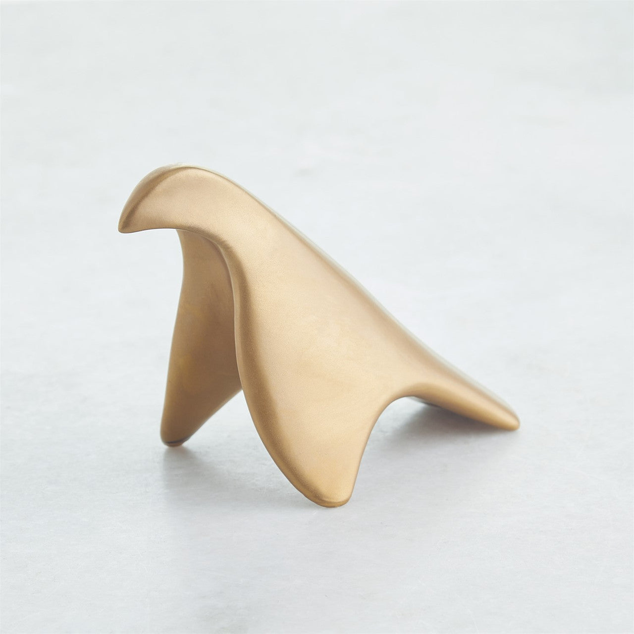Les Animaux Colombe Gold by Global Views is a smooth, abstract gold sculpture reminiscent of a stylized bird, showcased against a plain, light gray background.