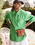 A smiling woman in a black cap, bright green shirt, and white skirt stands outdoors with her hands on her hips, carrying the Clare Vivier Crossbody Strap S25 bag. Patio furniture and greenery are visible in the background.