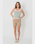 A person stands against a plain background, wearing a pink tank top with green stripes, The City Short by Kule made in Italy from cotton, and white sneakers. Their hands are on their hips.