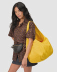 A woman with long black hair wears a brown patterned shirt, black skirt, and black woven belt bag. She carries Clare Vivier’s Grand Bando Canvas tote in bright yellow with external pockets against a plain light background.
