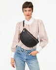 A woman with short brown hair wears a striped blouse, blue jeans, and carries the Grande Fanny by Clare Vivier—a black crossbody bag with an adjustable strap—while standing against a plain white background.