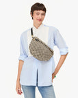 A smiling person in a light blue button-up shirt and jeans carries the Clare Vivier Grande Fanny S25, a black-and-white checkered crossbody bag with an adjustable strap, while standing against a plain white background.