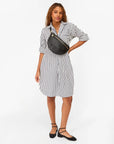 A woman with long, wavy hair poses against a white background, wearing a white shirt dress with black stripes, black flats, and the Grande Fanny S25 by Clare Vivier—a handwoven leather crossbody bag with an adjustable strap.