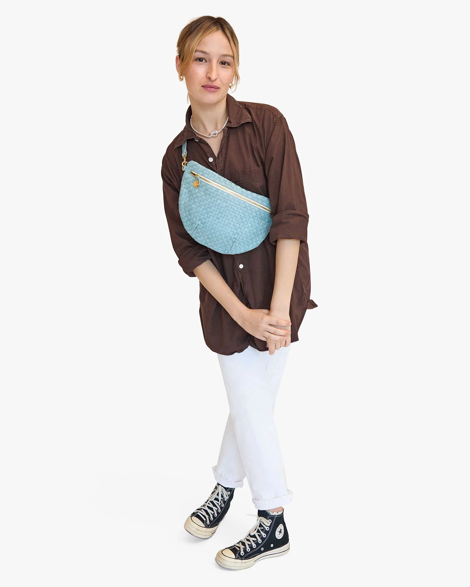 A young woman in a casual outfit, with a brown shirt, white pants, and black sneakers, poses with a Clare Vivier Grande Fanny Woven Checker featuring an adjustable strap, standing against a plain white background.