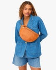 A woman with long braids wears a blue denim jacket and shorts against a white background, carrying the Clare Vivier Grande Fanny S25—a large handwoven leather crossbody bag with adjustable strap—slung across her chest.
