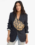 A woman with long dark hair wears a dark denim blazer, light wash jeans, and a chunky gold necklace. She carries the Clare Vivier Grande Fanny S26, a tan and black handwoven leather crossbody bag, across her chest.