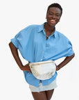 A smiling woman stands with her hands on her hips against a white background, wearing a loose blue button-up shirt, light denim shorts, a chunky necklace, and the Clare Vivier Grande Fanny S25 handwoven leather bag with adjustable strap.