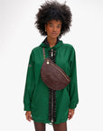 A woman with curly hair, wearing a green hooded jacket with black trim, carries the Clare Vivier Grande Fanny S26—a large handwoven leather bag with an adjustable strap—against a plain white background.