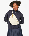A smiling person with curly hair wears a navy button-up shirt, a pleated skirt, and carries the Clare Vivier Grande Fanny S25, a handwoven leather crossbody bag with an adjustable strap and wooden charms.
