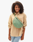 A person with curly hair smiles in a beige button-up shirt and light blue pants, carrying the Grande Fanny S25 by Clare Vivier—a green and white checkered crossbody bag with a detachable strap—over their shoulder. The background is plain white.