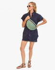 A woman in a navy shirt dress, denim shorts, and sandals stands holding the Clare Vivier Grande Fanny S25—a green patterned crossbody bag with an adjustable strap. She wears sunglasses and looks to the side with one hand on her hip.