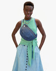 A person stands with hands in pockets wearing a green and white striped sleeveless top, a denim skirt with front buttons, and carries the Clare Vivier Grande Fanny S25 crossbody bag; a shirt is tied around their waist against a plain background.