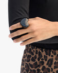 A person in leopard-print pants and a black top rests their left hand on their right arm, showcasing the Grande Heart Ring / Black by Clare Vivier (size 7) on their index finger—a chic accessory that completes the look.