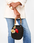 A person in a cream jacket and blue jeans carries a black woven handbag with a bamboo handle, adorned with Clare Vivier’s Grand Mylar Heart FOB charm and gold and red accents. They wear a large round ring on their hand.