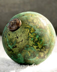 The Creative Co-op Marbled Glass Ornament showcases a green, patina-inspired finish with dark green and brown streaks and a metallic cap, resting elegantly on snowy surfaces.