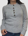 A person wears the White + Warren Cashmere Luxe Henley in light gray, featuring five brown buttons diagonally from collarbone to chest, paired with dark blue jeans against a plain white background.