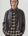 A woman with straight dark hair wears the SUNCOO LENNOX black pleather jacket over a black and beige plaid button-up shirt, paired with dark trousers and gold hoop earrings, standing against a plain light background.