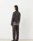 A person with straight, dark hair faces away, wearing the LENNOX Jacket by SUNCOO in dark brown pleather and matching pants. A plaid shirt peeks out beneath the jacket against a plain white background.