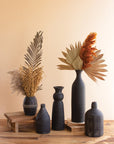 Five Kalalou, Inc Black Clay Vases with dried pampas grass, palm leaves, and botanicals are displayed on a wooden riser and surface against a beige wall, adding a minimalist rustic accent to modern home decor.
