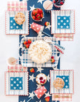 A festive summer barbeque table with My Mind's Eye Cocktail Paper Napkins, star-spangled plates, a whipped cream pie, mini pies, berries, pink drinks, and red, white & blue decor for a holiday celebration.