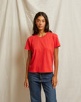 A person with long dark hair stands against a beige fabric backdrop, wearing the Perfectwhitetee Harley SS Boxy Crew in red and mid-rise denim jeans, looking at the camera with a neutral expression.
