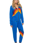 A woman with long, wavy blonde hair and a neutral expression stands barefoot wearing Aviator Nation's all seasons circle chevron women's sweatpant, styled in a bright blue with vintage-inspired chevron stripes.