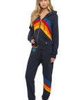 A woman with long blonde hair and a neutral expression stands barefoot in Aviator Nation’s all seasons circle chevron women’s sweatpants, featuring bold diagonal stripes for a vintage vibe, styled casually with a matching zip-up hoodie.
