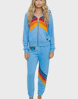 A woman with long wavy blonde hair models Aviator Nation’s all seasons circle chevron women’s sweatpant, featuring bold orange, red, and navy stripes. She wears a matching light blue zip-up hoodie and stands barefoot against a plain white background.