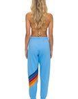 A woman with long, wavy blonde hair stands barefoot, facing away. She wears a tan bikini top and Aviator Nation's all seasons circle chevron women's sweatpant, light blue jogger style with multicolored stripes, handmade in Los Angeles.