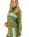 A woman with long, wavy blonde hair wears the Aviator Nation 5 stripe cropped pullover hoodie relaxed in green camo with blue and yellow stripes. She stands facing forward, one hand in her pocket, looking at the camera.