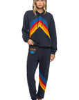 A woman with loose hair and a neutral expression stands barefoot wearing the Aviator Nation mountain stitch chevron pullover hoodie relaxed and matching sweatpants, featuring colorful chevron stripes in red, orange, yellow, and blue.