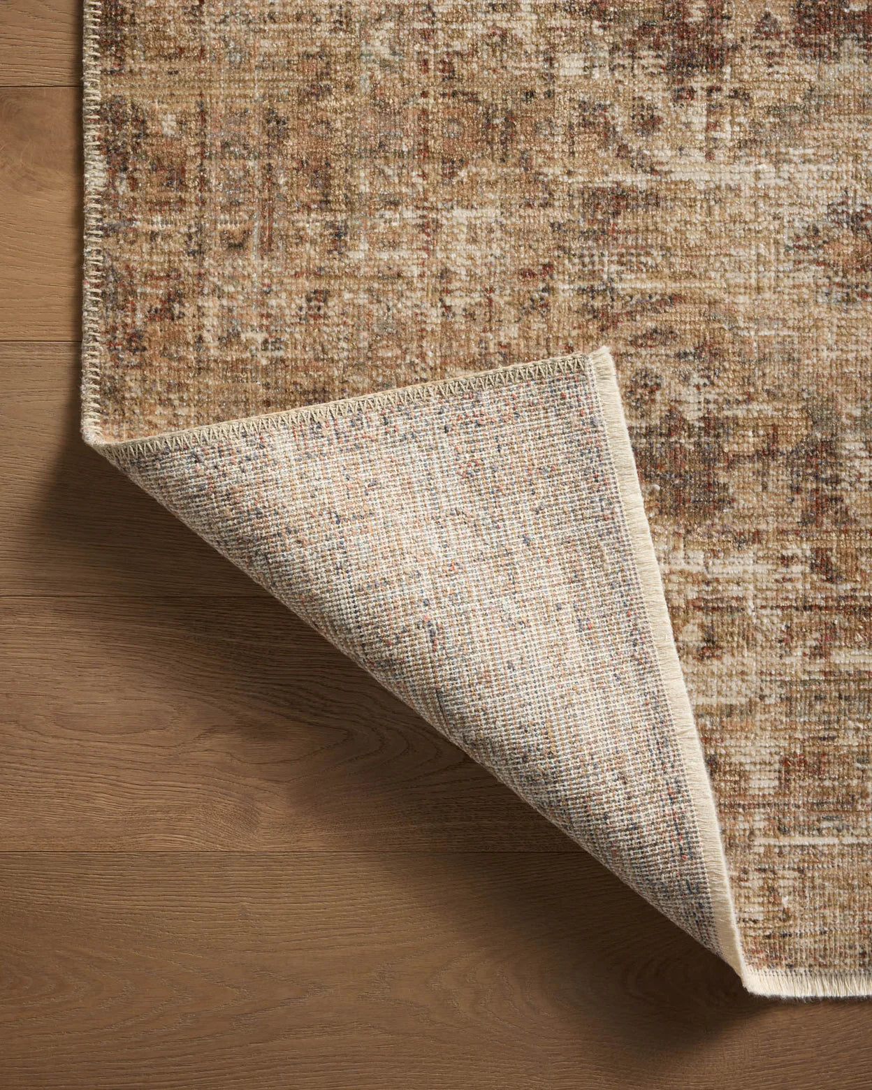 A close-up of a textured Loloi Rugs Bark / Multi Rug with warm, Arizona-inspired tones, partially rolled up to reveal its underside, on a wooden floor.