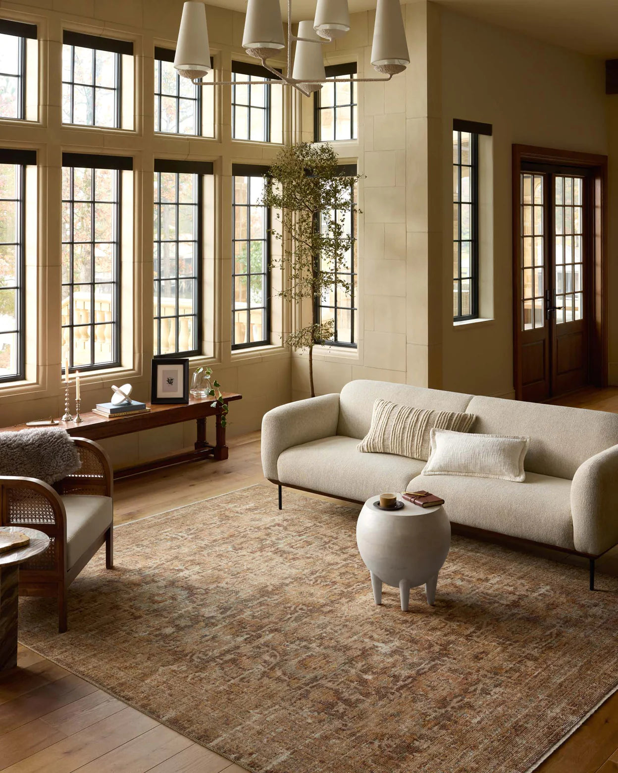An elegant living room with large windows, beige walls, and a classic cream sofa. A small, rounded table and a wicker chair add to the cozy, well-lit space in Arizona style with a Bark / Multi Rug by Loloi Rugs.