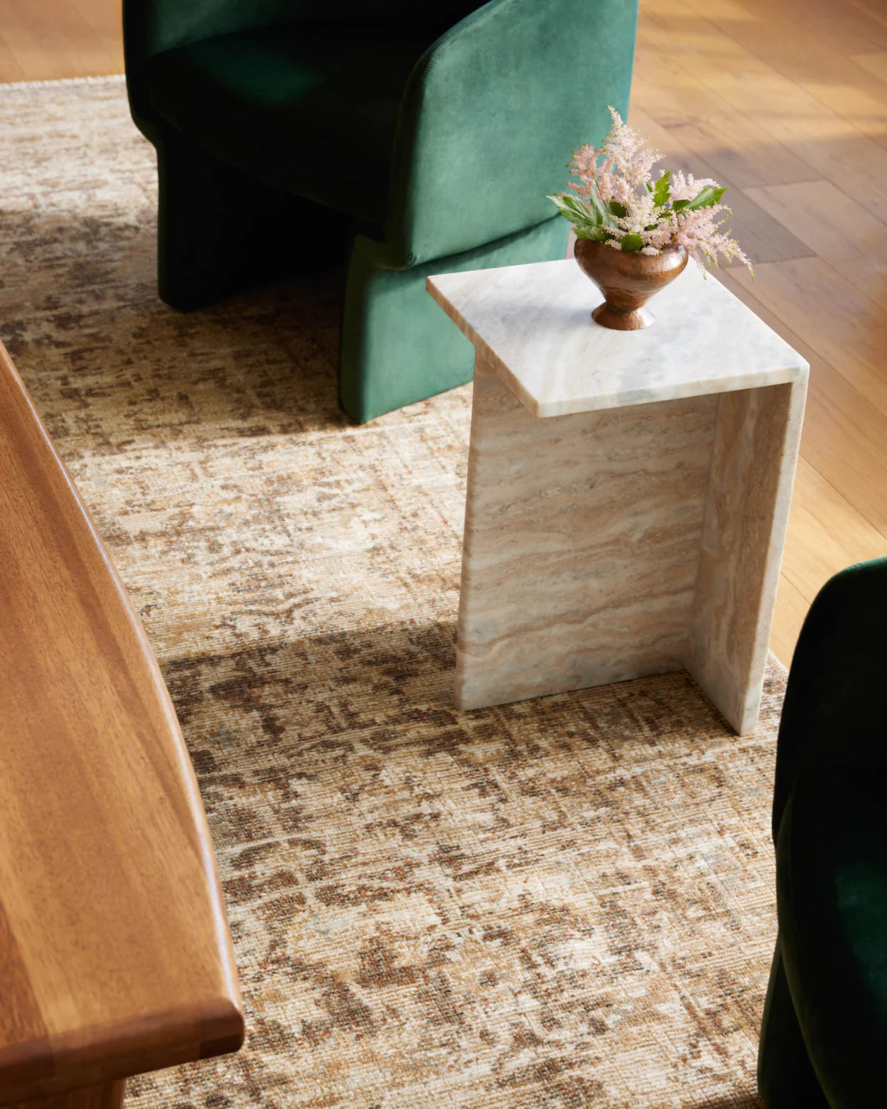 A stylish interior with a modern marble side table holding a small vase of flowers, positioned on a Loloi Rugs Brick / Multi Rug beside a green velvet chair and a wooden table in a Scottsdale Arizona bungalow.