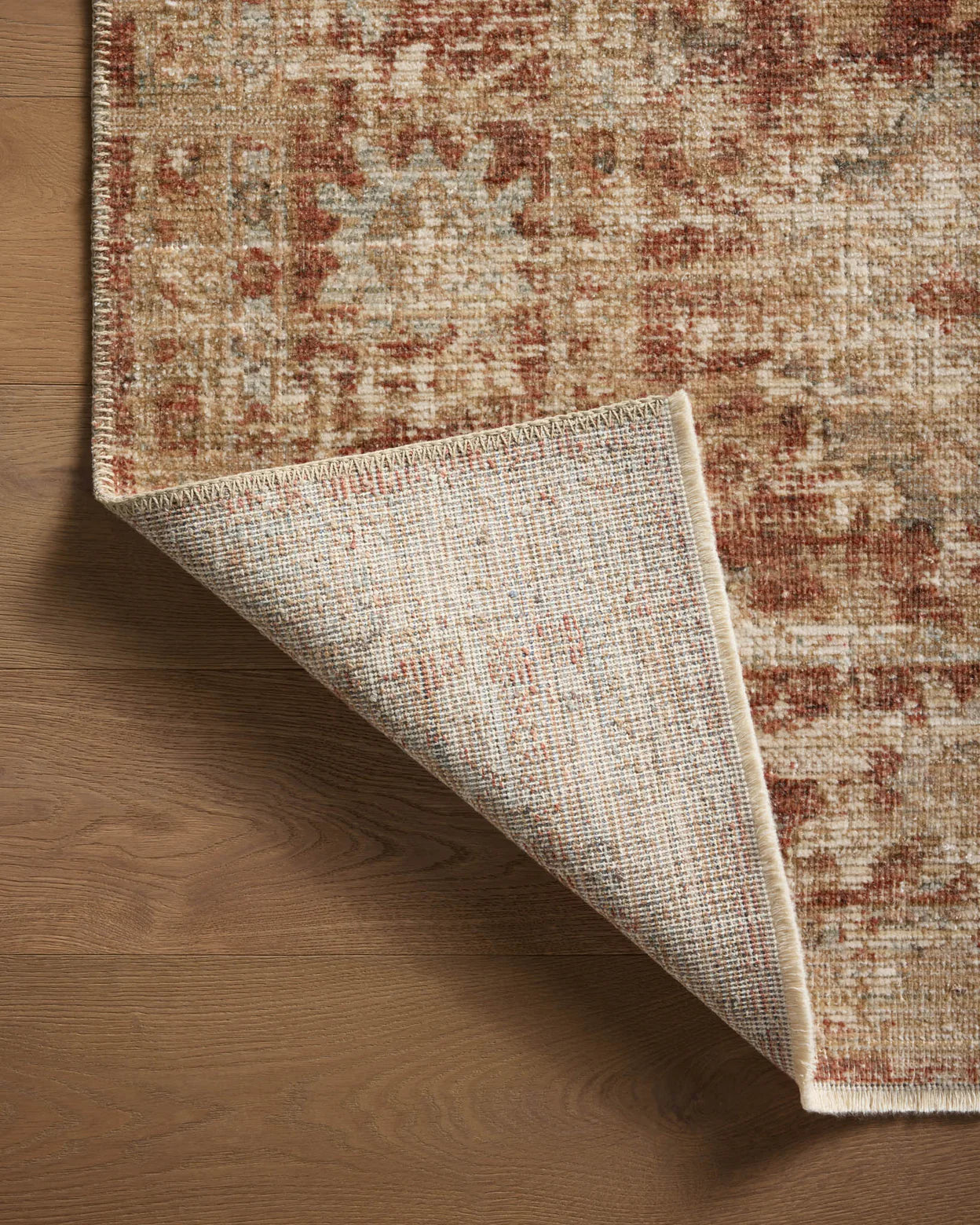 A detailed view of a corner of an ornate, patterned Loloi Rugs Brick / Multi Rug partially laid over a smooth wooden floor in a Scottsdale Arizona bungalow, with the underside of the rug visible as it's slightly folded back.
