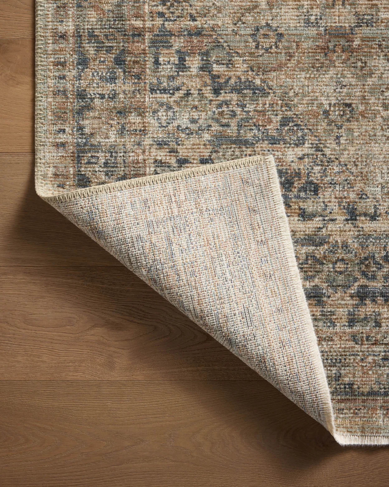 A corner of a decorative area rug in a Scottsdale bungalow is flipped back to reveal its underside on a wooden floor, showcasing a detailed, multicolored pattern on the top surface of the Sage / Navy Rug by Loloi Rugs.