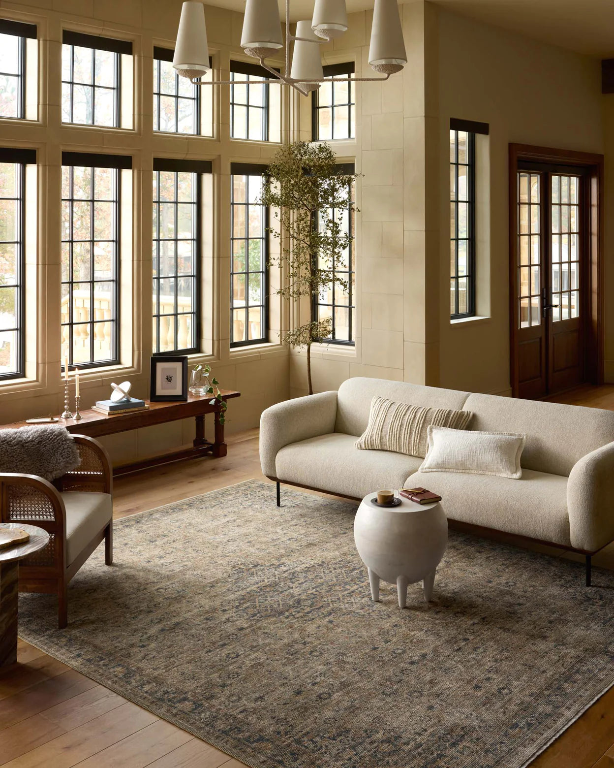 A cozy living room in a Scottsdale bungalow, featuring a large beige sofa, a white round coffee table, and a wicker chair. The room includes large windows, wooden floors, and beige walls decorated with touches of greenery. The room is anchored by a Sage / Navy rug from Loloi Rugs.