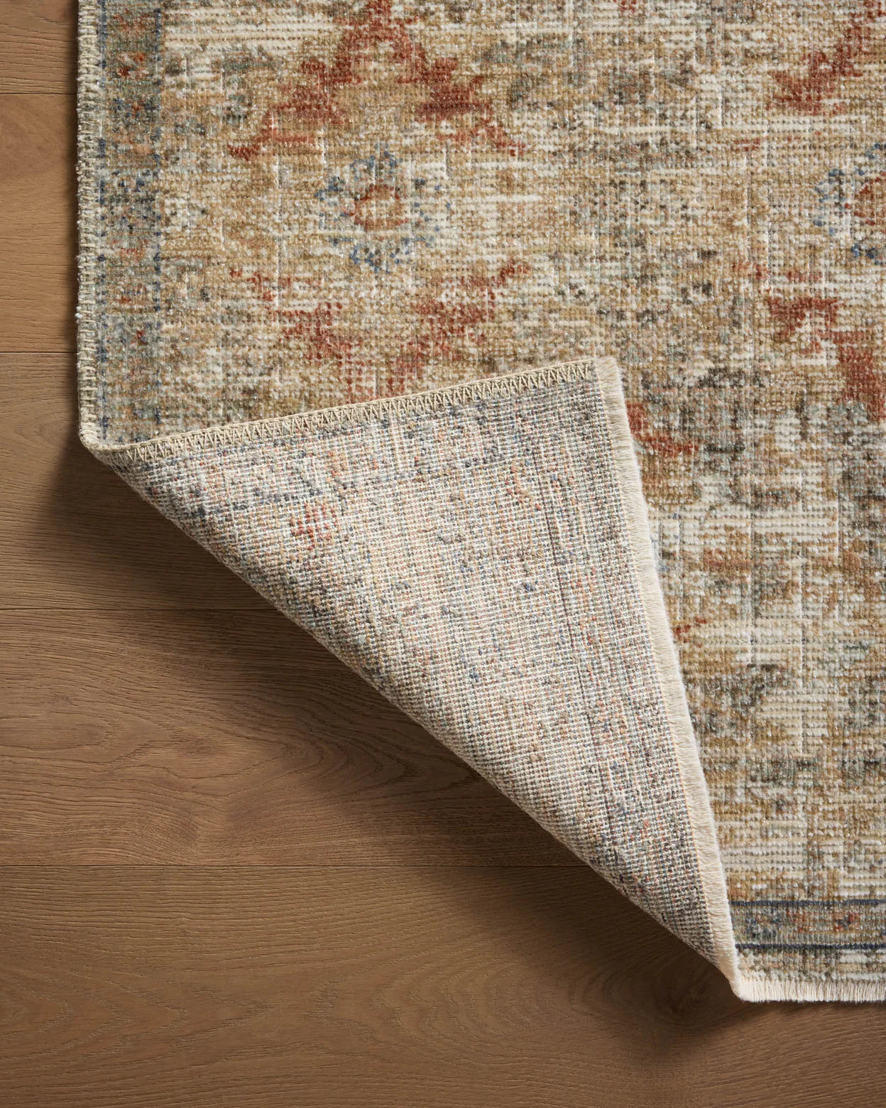 A corner of an ornate Loloi Rugs Grey / Sunset Rug in a Scottsdale bungalow, with a detailed, multicolored pattern, slightly lifted to reveal its texture, on a wooden floor.