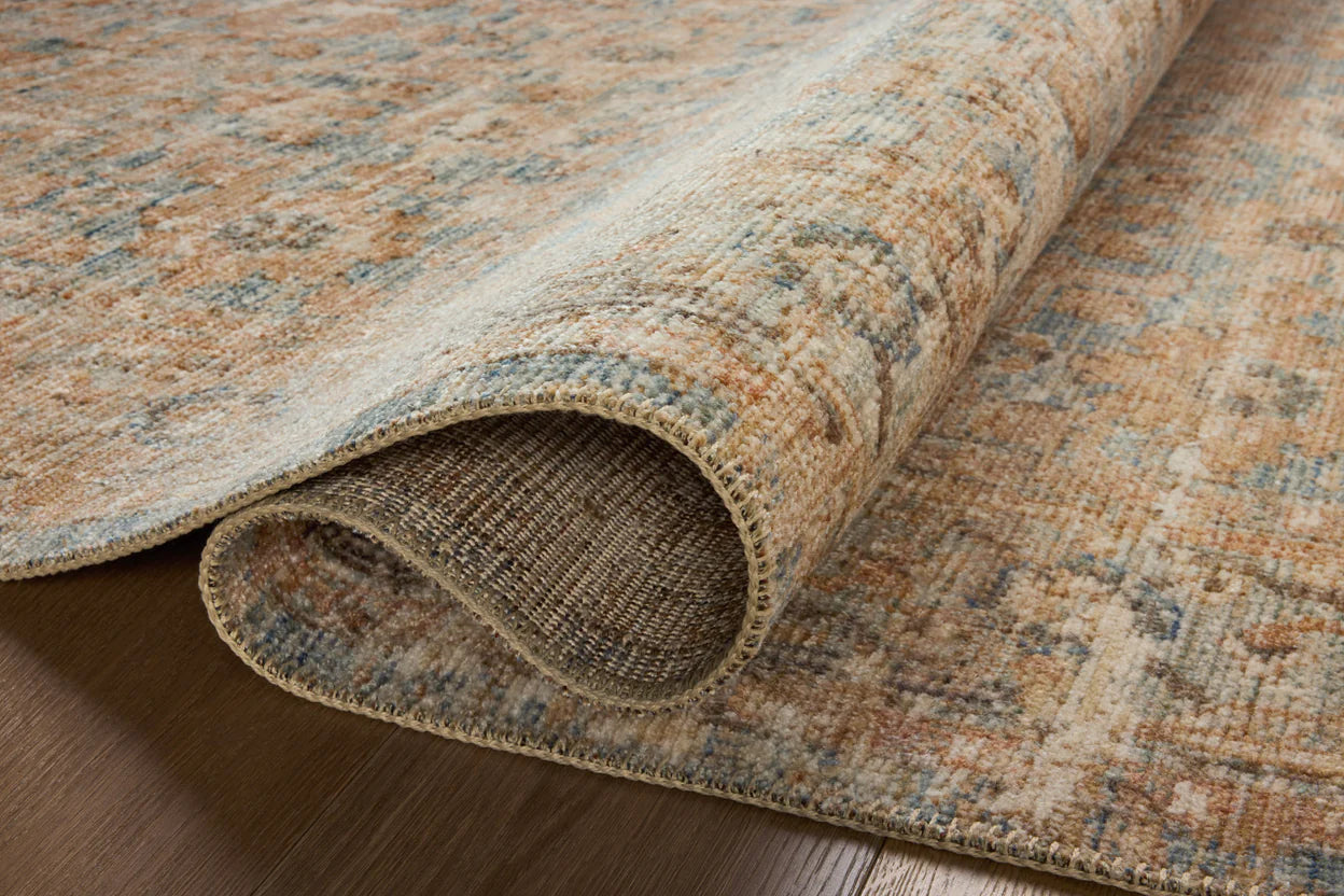 A close-up of a partially rolled-up Ocean / Sand Rug on a wooden floor in a Scottsdale, Arizona bungalow, displaying intricately faded patterns in earth tones from Loloi Rugs.