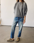 Indoors, a person wears Hey Gang's The Boxy Crop Sweatshirt in gray, blue jeans, and tan slip-on shoes. Their right hand rests in a pocket; their head is out of frame.