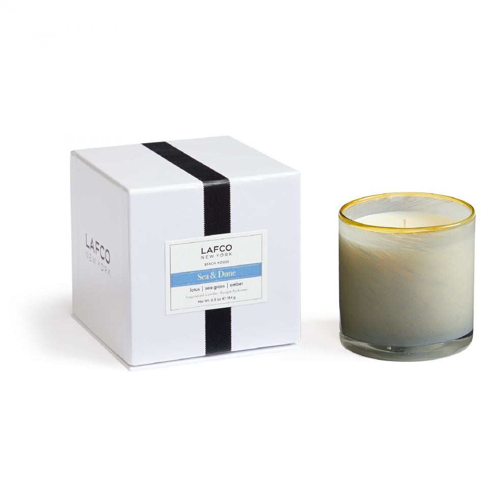 A Lafco NY Beach House Candle made of soy wax sits in a hand-blown glass vessel with a gold rim, next to its white box featuring a black stripe and blue scent label.